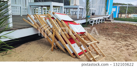 Neatly stacked red and white wooden lounge chairs on a sandy terrace next to a beach hut. Concept of the end of the beach season, a small business in operation Neatly stacked red and white wooden lounge chairs on a sandy terrace next to a beach hut. Concept of the end of the beach season, a small business in operation 132103622