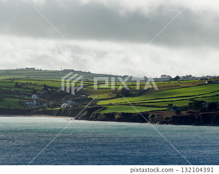 Beautiful landscape shows the coastline of Dunmore Bay Beach in Ireland. Green terraced hills slope down to the rocky coast and light waves under a gray cloudy sky. Beautiful landscape shows the coastline of Dunmore Bay Beach in Ireland. Green terraced hills slope down to the rocky coast and light waves under a gray cloudy sky. 132104391