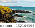 Waves crash against jagged rock formations along the coast. Hills are visible in the distance, creating a scenic landscape on a sunny day in Ireland. 132104394