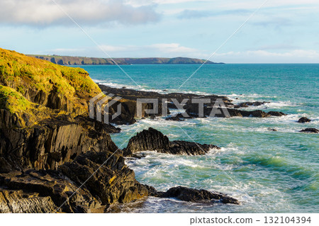 Waves crash against jagged rock formations along the coast. Hills are visible in the distance, creating a scenic landscape on a sunny day in Ireland. Waves crash against jagged rock formations along the coast. Hills are visible in the distance, creating a scenic landscape on a sunny day in Ireland. 132104394