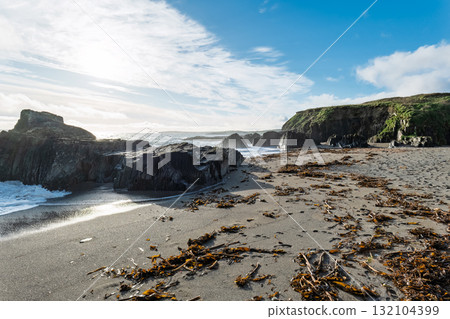 Coastal scene shows waves crashing on rocks at Sheep Cove, Ireland. The sandy beach is covered with seaweed. Blue sky and some clouds are overhead. 132104399