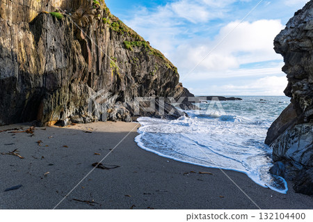 Waves crash onto the dark sand beach at Sheep Cove in Ireland, with steep, rocky cliffs rising up on either side under a partly cloudy, blue sky. Waves crash onto the dark sand beach at Sheep Cove in Ireland, with steep, rocky cliffs rising up on either side under a partly cloudy, blue sky. 132104400