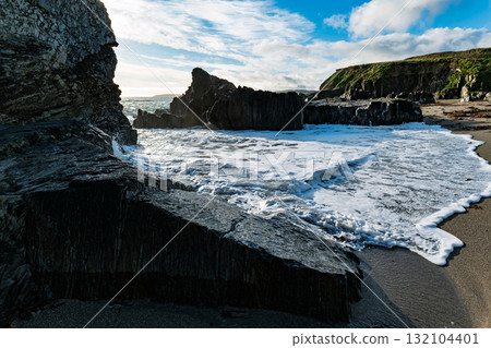 On a sunny day, waves from the ocean gently wash up on the black sand beach at Sheep Cove in Ireland, among the rocks and cliffs. On a sunny day, waves from the ocean gently wash up on the black sand beach at Sheep Cove in Ireland, among the rocks and cliffs. 132104401