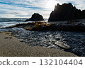A small creek flows onto the sandy beach and out into the ocean at Sheep Cove in Ireland. Seaweed is scattered on the beach near dark, jagged rock formations. 132104404
