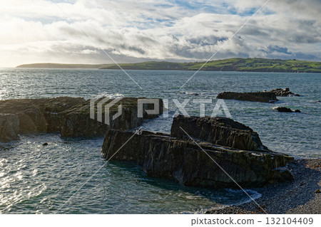 Rocky coast at Sheep Cove in Ireland shows the beautiful blue ocean and green hills. A cloudy sky hangs over the landscape and rocky formations extend into the water. 132104409