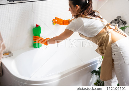 Woman in rubber gloves and apron manually washes bathtub is in bathroom of house with sponge cleaner and cleaning agent in a bottle. Restoring order, cleanliness and hygiene in home, cleaning servise 132104501
