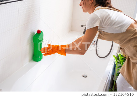 Woman in rubber gloves and apron manually washes bathtub is in bathroom of house with sponge cleaner and cleaning agent in a bottle. Restoring order, cleanliness and hygiene in home, cleaning servise 132104510