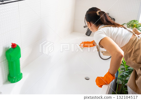 Woman in rubber gloves and apron manually washes bathtub is in bathroom of house with sponge cleaner and cleaning agent in a bottle. Restoring order, cleanliness and hygiene in home, cleaning servise 132104517