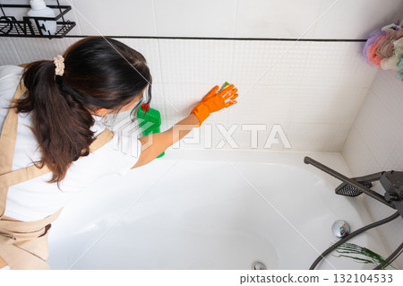 Woman in rubber gloves and apron manually washes bathtub is in bathroom of house with sponge cleaner and cleaning agent in a bottle. Restoring order, cleanliness and hygiene in home, cleaning servise 132104533