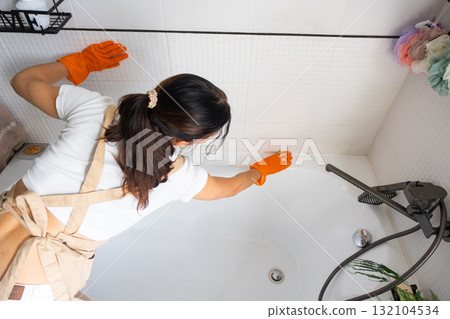 Woman in rubber gloves and apron manually washes bathtub is in bathroom of house with sponge cleaner and cleaning agent in a bottle. Restoring order, cleanliness and hygiene in home, cleaning servise 132104534