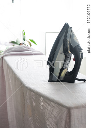 The iron is on the ironing board with a plain cloth in the room in close-up. The iron is on the ironing board with a plain cloth in the room in close-up. 132104732
