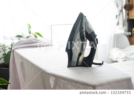 The iron is on the ironing board with a plain cloth in the room in close-up. 132104733
