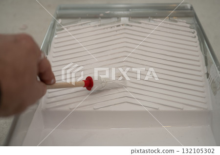 hand using a small round brush inside a plastic paint tray filled with white interior paint hand using a small round brush inside a plastic paint tray filled with white interior paint 132105302
