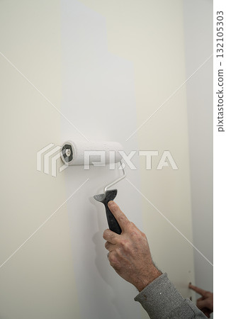 person painting an interior wall with a foam roller, applying a fresh white coat over a beige surface 132105303