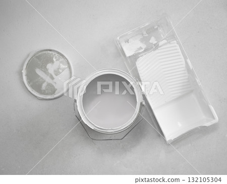 Top-down flat lay of an open paint can with white interior paint beside a plastic roller tray and lid on a neutral floor. Top-down flat lay of an open paint can with white interior paint beside a plastic roller tray and lid on a neutral floor. 132105304