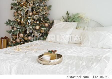 Christmas gift and cup on tray with lights on cozy white bed near decorated festive Christmas tree 132106949