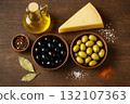 Top view of Mediterranean ingredients with cheese wedge, olive oil bottle, bowls of black and green olives, peppercorns, bay leaves, salt, and paprika on rustic wooden surface with copy space 132107363
