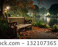 Night park scene with wooden bench on paved path surrounded by autumn leaves facing calm water reflecting streetlights and trees in misty atmosphere with copy space 132107365