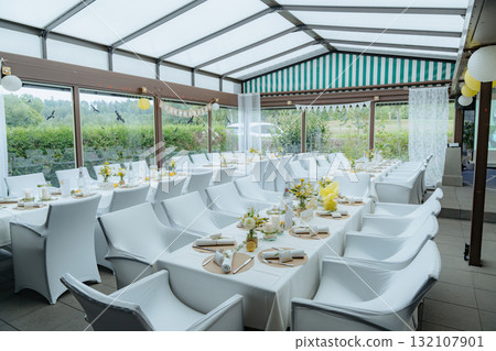 An elegantly decorated wedding reception in a glass-paneled venue with white chairs and tables set for guests. The setting features lush greenery outside, creating a serene atmosphere 132107901