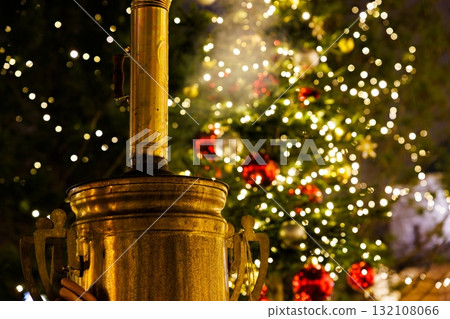 Golden kettle stands in the foreground with a beautifully lit Christmas tree behind at a festive location Golden kettle stands in the foreground with a beautifully lit Christmas tree behind at a festive location 132108066