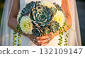 A hyper-realistic, high-detail close-up photograph of a bride's hands gently holding a gorgeous wedding bouquet filled with Large Succulents, cream Dahlias, trailing Sedum 132109147
