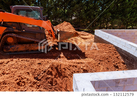 Skid steer loader is moving dirt at construction site next to foundation during works daytime. 132112579