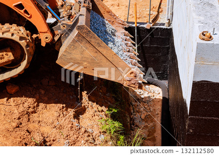 Excavator scoops gravel fills trench near building foundation during construction day Excavator scoops gravel fills trench near building foundation during construction day 132112580