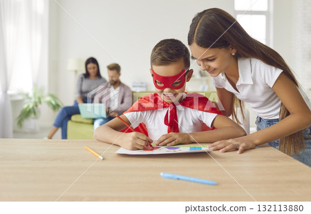 Happy little brother in superhero costume drawing pictures together with sister at home Happy little brother in superhero costume drawing pictures together with sister at home 132113880