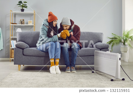 Man trying to warm his girlfriend's hand sitting on the sofa in winter coats near electric heater. Man trying to warm his girlfriend's hand sitting on the sofa in winter coats near electric heater. 132115931