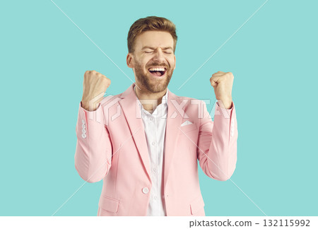 Young overjoyed happy man in pink suit with yes winner gesture and closed eyes on blue background. 132115992