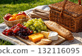 Picnic setup on grass with fruits, bread, cheese, and grapes beside a wicker basket 132116095