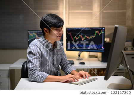 Young Asian trader analyzing financial charts on computer at night, focused on investment strategy 132117860