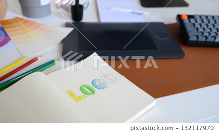 Close-up of logo sketch with color pencils, palette, and tablet on designer workspace in modern creative office 132117970