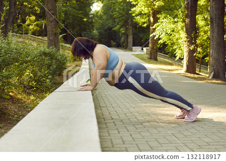 Motivated fat young woman working out doing push-ups in city park on summer morning. Motivated fat young woman working out doing push-ups in city park on summer morning. 132118917