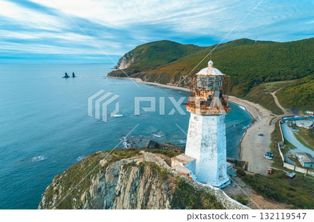 Lighthouse stands on rocky coastline, overlooking the ocean with mountains in the background during a clear day Lighthouse stands on rocky coastline, overlooking the ocean with mountains in the background during a clear day 132119547