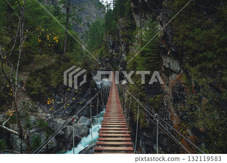 Wooden suspension bridge stretches over a rocky gorge surrounded by lush green trees in the mountains 132119583