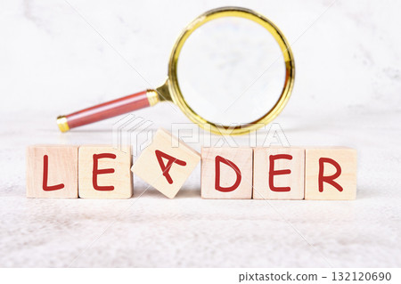 Wooden blocks arranged to spell leader sit on a light background, accompanied by a magnifying glass that symbolizes the search for leadership qualities and attributes 132120690