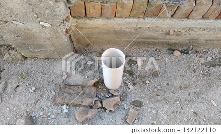 A white pvc pipe stands upright on a rough ground surface near a brick and concrete structure edge 132122110
