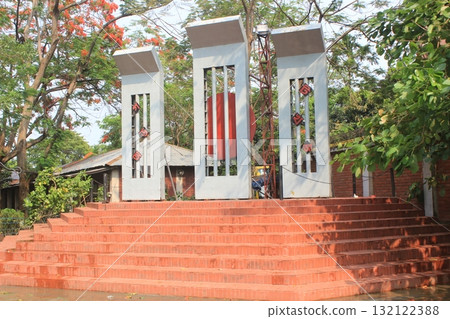 A striking modern monument with three tall white pillars stands on a wide red brick staircase, surrounded by lush green trees A striking modern monument with three tall white pillars stands on a wide red brick staircase, surrounded by lush green trees 132122388