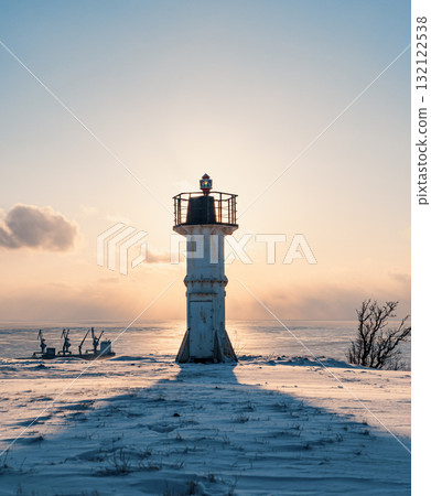 A white lighthouse stands tall on a snowy shore, casting a long shadow as the sun sets over the frozen sea 132122538