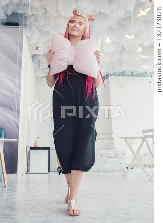 A young woman poses with a large bow-shaped pillow, showcasing her vibrant pink and blonde braids. 132123028