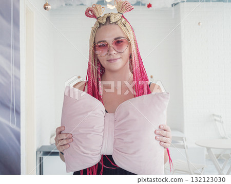 A young woman poses with a large bow-shaped pillow, showcasing her vibrant pink and blonde braids. 132123030