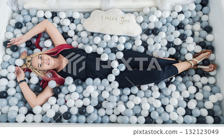 Young woman relaxes in a playful ball pit filled with monochrome spheres and a cloud pillow 132123049