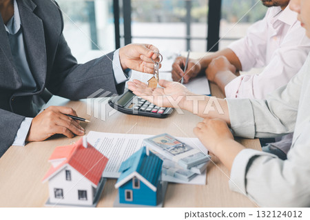 Close up of real estate agent handing house key to client with contract paperwork, calculator and house models on desk, concept of home ownership, mortgage loan, property investment and success. Close up of real estate agent handing house key to client with contract paperwork, calculator and house models on desk, concept of home ownership, mortgage loan, property investment and success. 132124012