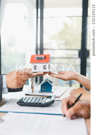 Close up of real estate agent handing house model to client with calculator and contract paperwork on desk, concept of mortgage loan, home ownership, property investment and housing agreement. Close up of real estate agent handing house model to client with calculator and contract paperwork on desk, concept of mortgage loan, home ownership, property investment and housing agreement. 132124113