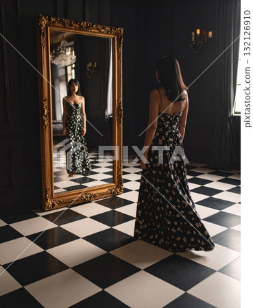 Woman in an evening dress standing on a checkered floor and looking into a mirror with a strange reflexion, conceptual horror scene Woman in an evening dress standing on a checkered floor and looking into a mirror with a strange reflexion, conceptual horror scene 132126910