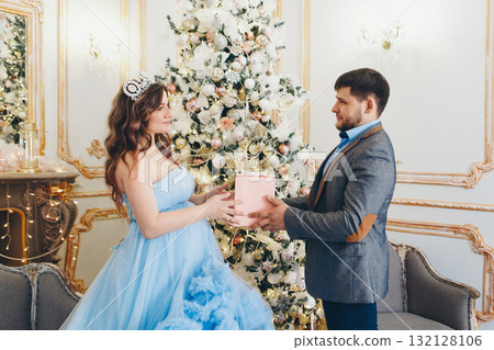 husband in a gray suit gives a gift to his pregnant wife. pregnant girl in a lush blue dress. young man in a blue shirt. married couple standing by the Christmas tree. family photo session.  132128106
