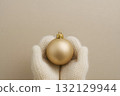 Hands in cream knitted mittens holding a matte gold Christmas bauble on a beige background 132129944