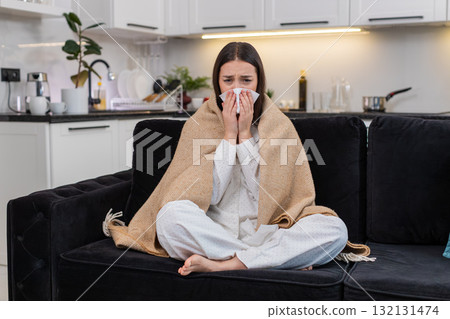 Young woman wrapped in blanket sneezes uses tissue cold flu symptoms sniffles unwell sick tired weak 132131474