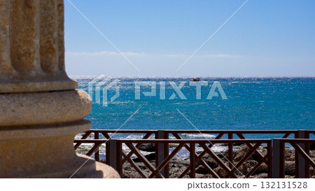 Scenic View of Calm Azure Waters with Sailboat on the Horizon and Breezy Coastal Environment Framed by Architectural Pillar Scenic View of Calm Azure Waters with Sailboat on the Horizon and Breezy Coastal Environment Framed by Architectural Pillar 132131528